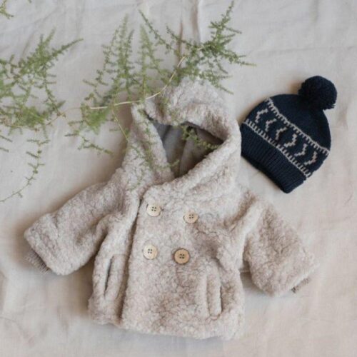 Giacca in pelliccia sintetica beige per bambini con cappuccio e bottoni, abbinata a un cappellino blu in maglia.