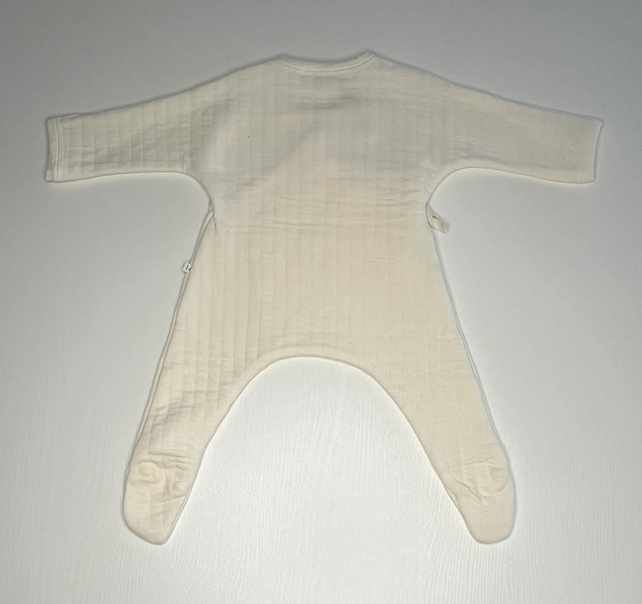Tuta per neonati in tessuto chiaro con chiusura incrociata e fiocco laterale.